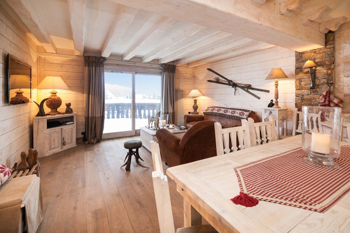 Rustic ski chalet interior featuring wood-paneled walls, beamed ceilings, a dining area, and a living space with a large window opening to a snowy mountain vista.