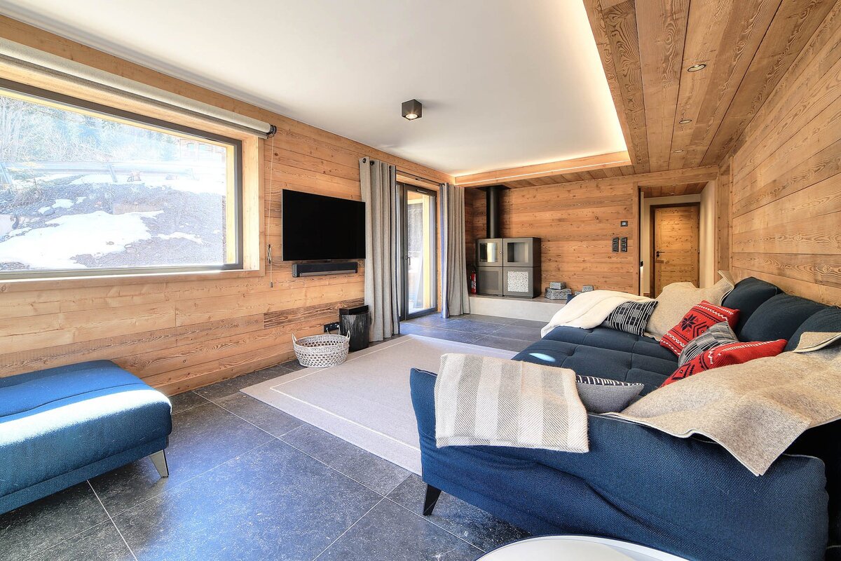 Cozy chalet living room with warm wood paneling, a comfy blue sofa, flat-screen TV, large window overlooking snow, and a modern wood-burning stove.