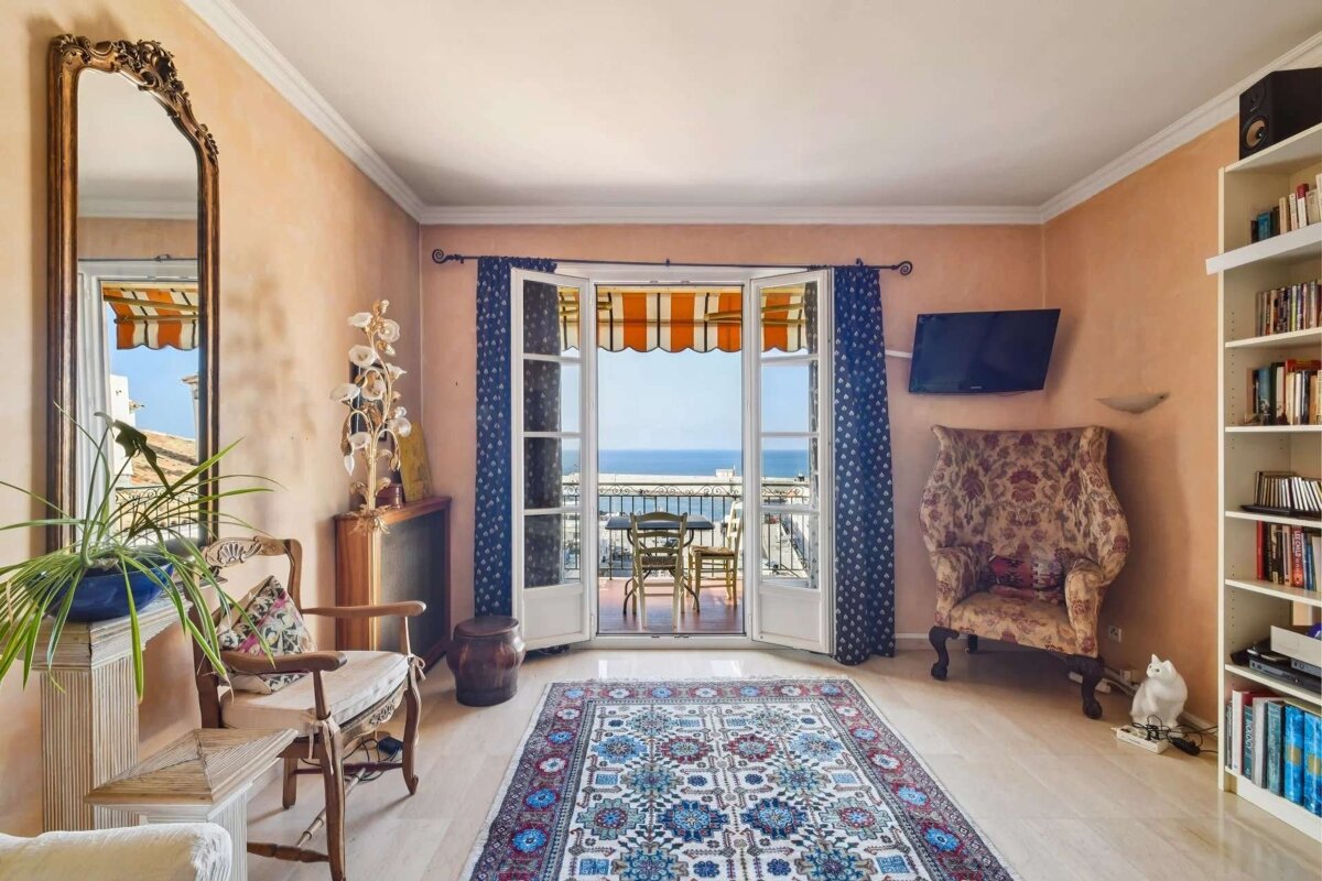 A bright, peach-walled room with a balcony offering a stunning sea view. Features include a colorful rug, an ornate mirror, and a full bookshelf.