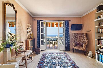 A bright, peach-walled room with a balcony offering a stunning sea view. Features include a colorful rug, an ornate mirror, and a full bookshelf.