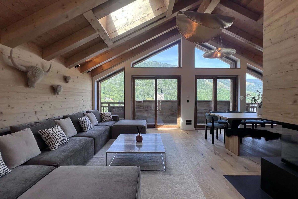Bright chalet living room with wood interior, vaulted ceiling, and large windows revealing mountain views. Modern grey sofa and dining area.