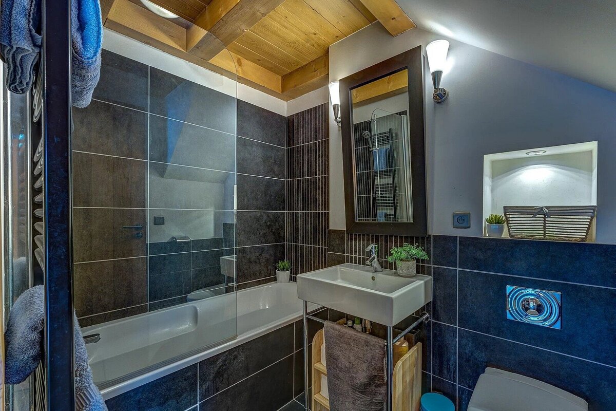 Modern bathroom with a wooden ceiling, dark tile, a bathtub with a glass screen, a sink, and a mirror.