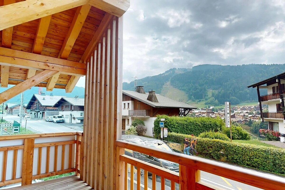 View from a wooden chalet balcony of an Alpine village with traditional homes and forested mountains under a cloudy sky.