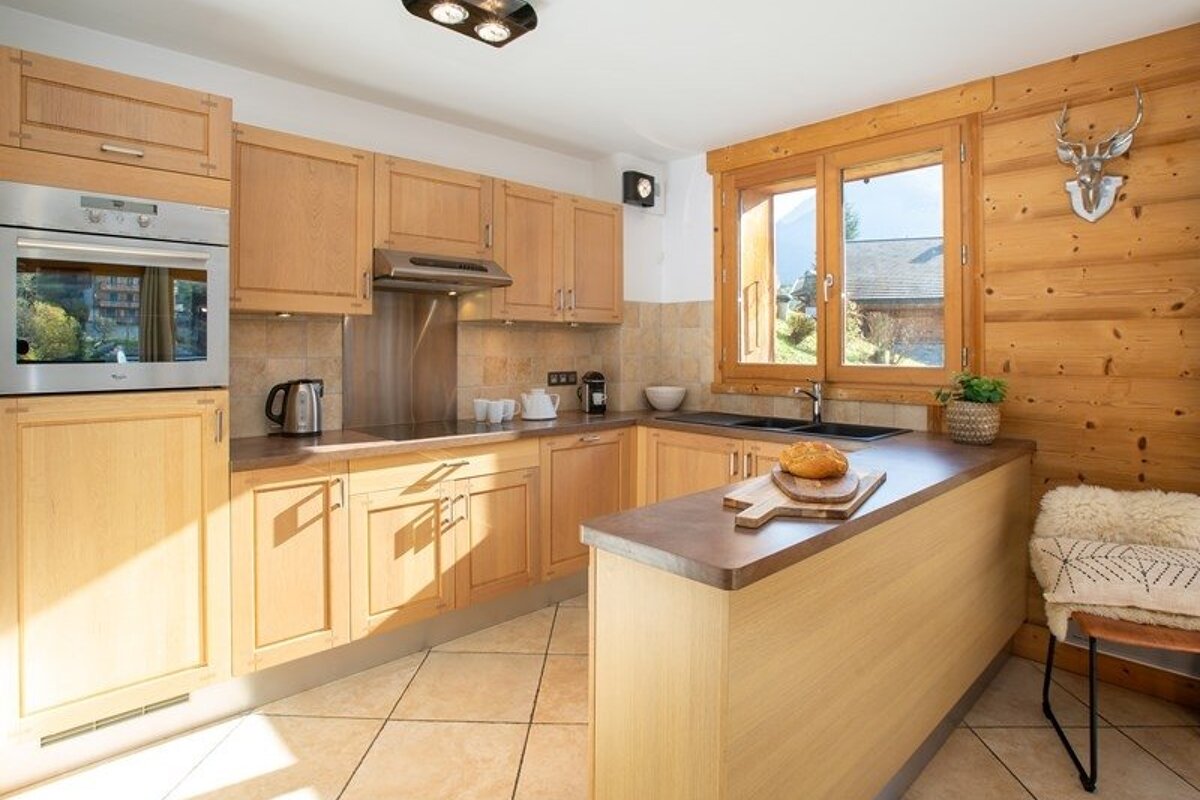 A rustic kitchen featuring light wood cabinets, tiled flooring, a wooden paneled wall, and a breakfast bar with a loaf of bread.