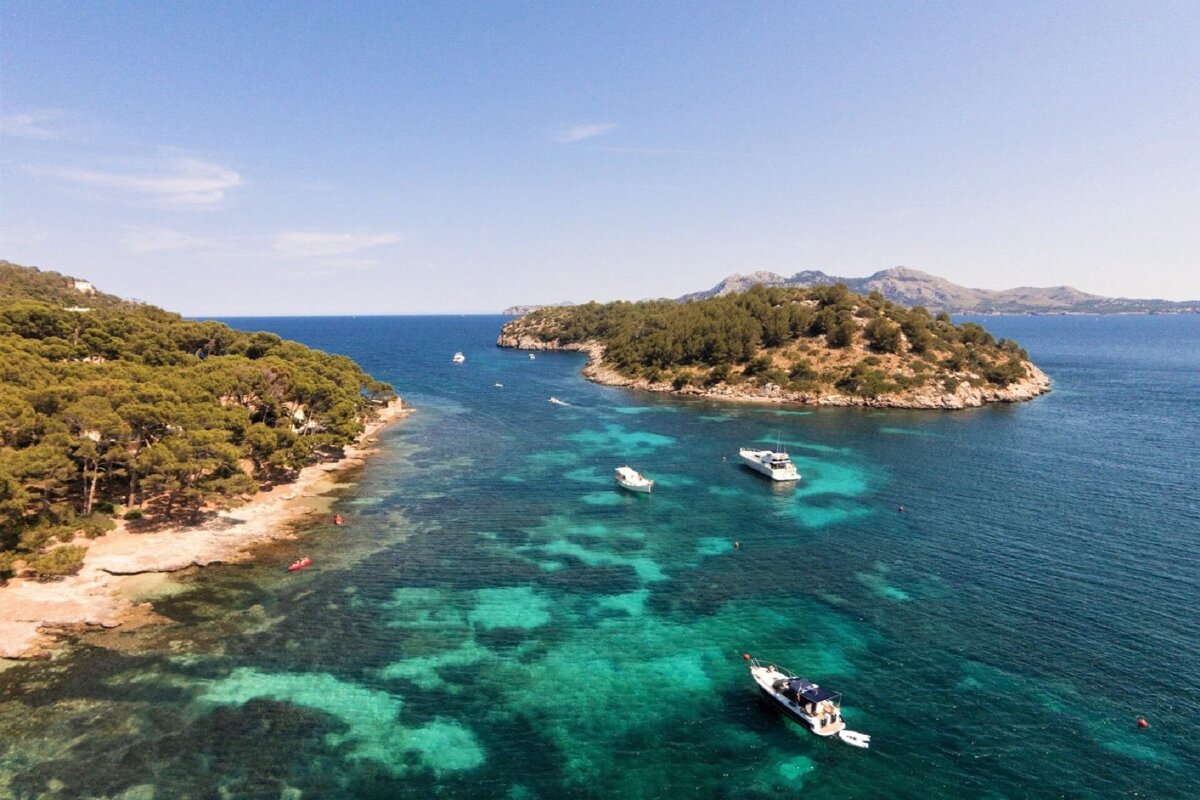 An aerial view of a stunning turquoise bay with multiple boats anchored near lush, tree-covered coastlines and distant mountains under a clear sky.