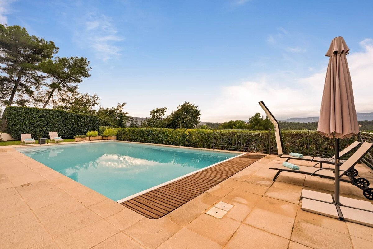 A sunny patio featuring a blue swimming pool, lounge chairs under an umbrella, and green trees against a clear blue sky.