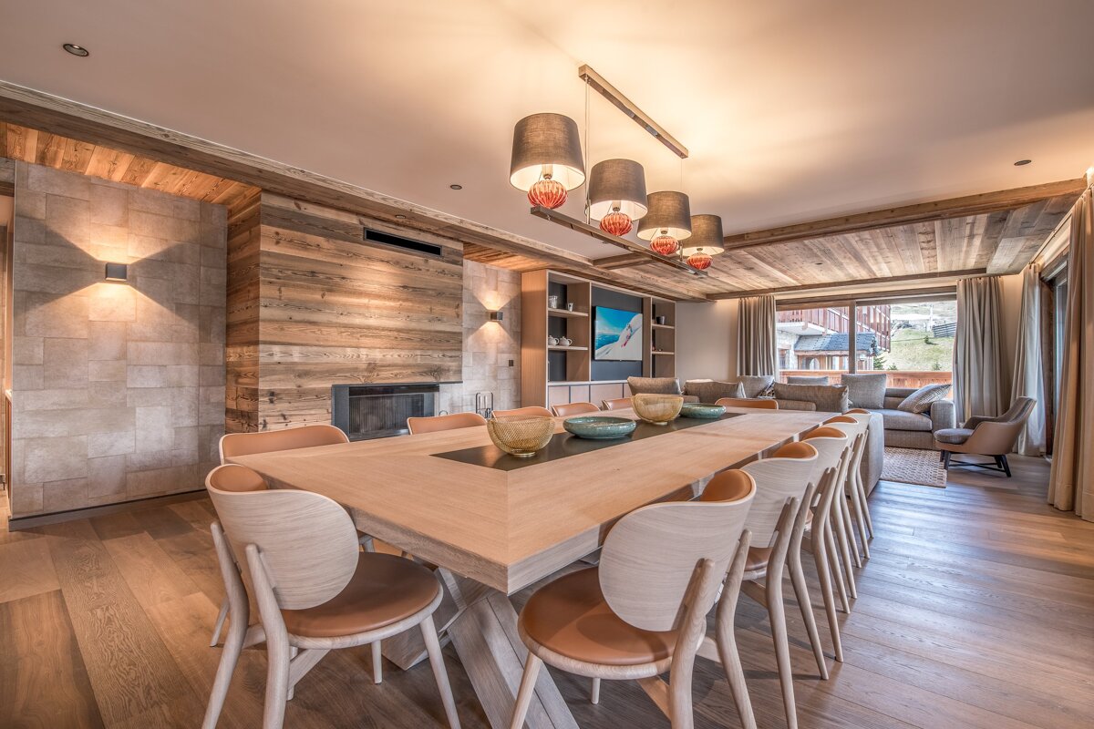 A chic, warm chalet-style open-plan room with a large wooden dining table, fireplace, and living area. Features textured wood walls & ceiling, stylish lighting.