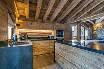 A rustic modern kitchen featuring rich wooden walls, cabinets, and exposed ceiling beams, complemented by dark countertops and modern appliances.