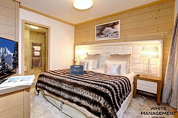 A luxurious, cozy bedroom featuring a large bed with a furry throw, wood-paneled wall, TV displaying mountains, and an en-suite bathroom.