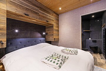 Rustic bedroom with wooden cabinetry, a white-dressed bed, and an adjoining modern bathroom with dark grey tiles.