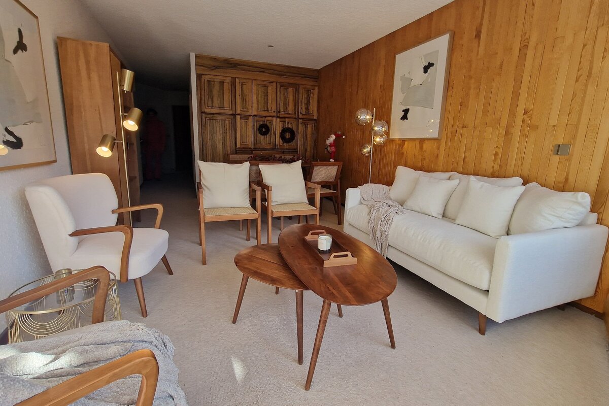 A cozy mid-century modern living room featuring a white sofa, armchairs, wooden coffee tables, and wood-paneled walls with warm lighting.
