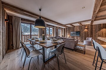 A luxurious chalet interior featuring a dining table set for guests, a cozy living area, and large windows revealing a snowy mountain view, all in warm wood tones.