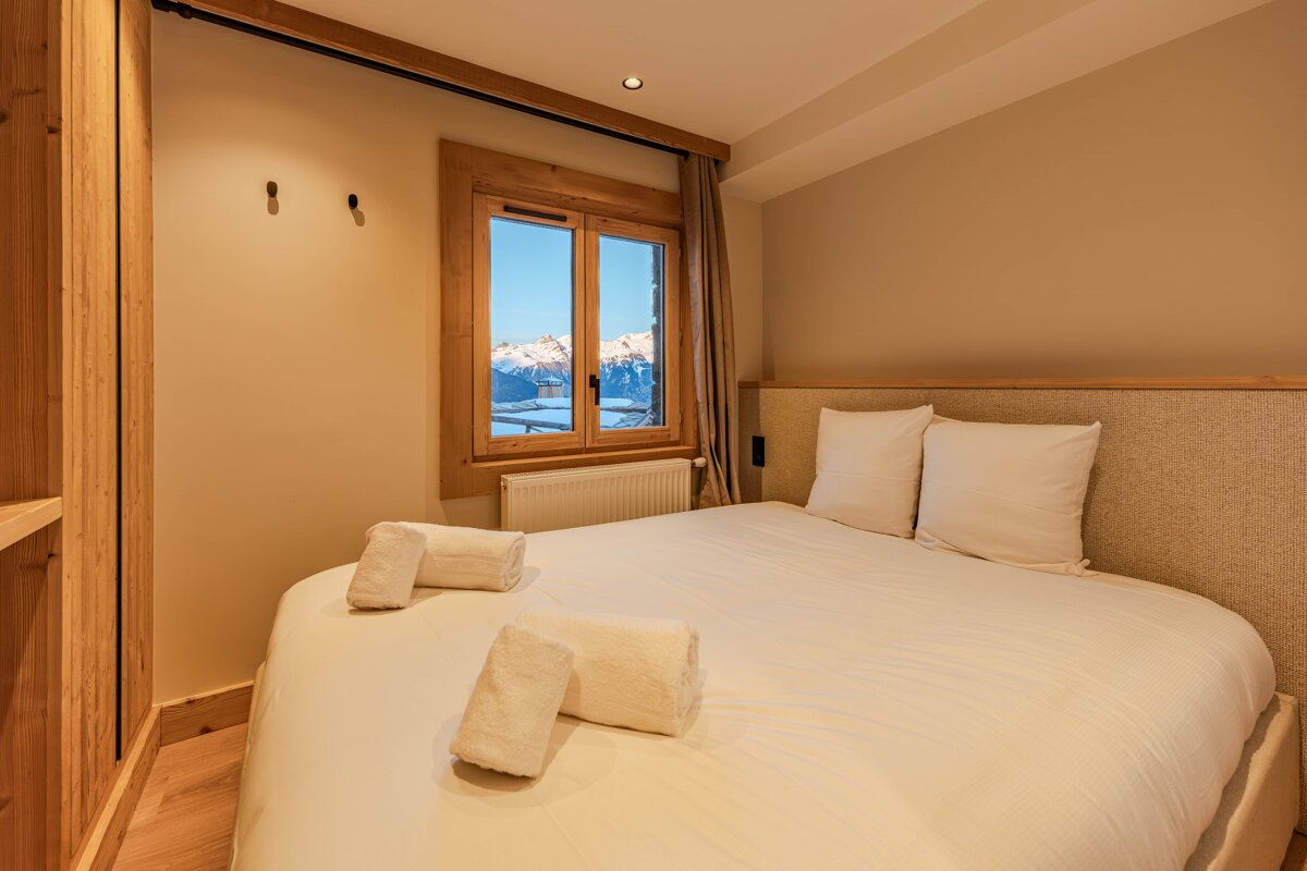 Cozy chalet bedroom featuring a bed with white linens and a window looking out onto snowy mountains.
