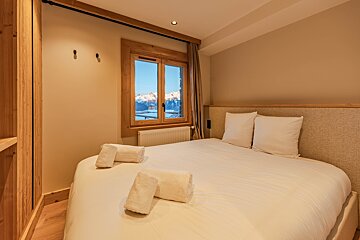 Cozy chalet bedroom featuring a bed with white linens and a window looking out onto snowy mountains.