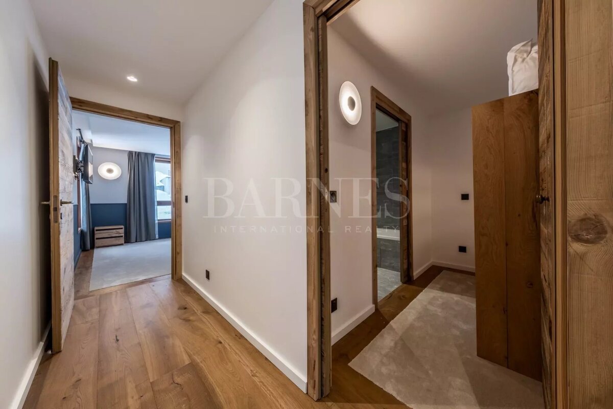 A stylish interior hallway featuring light wooden flooring, white walls, and natural wood door frames. Doors lead to a bedroom and another room, with modern lighting.