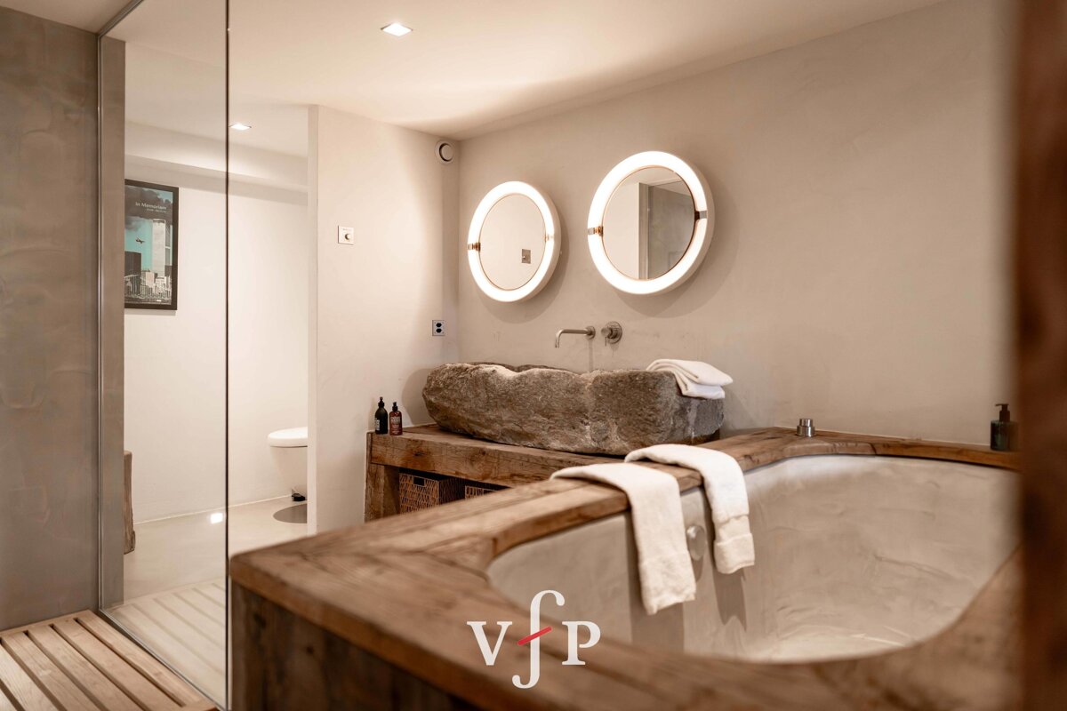 A chic rustic bathroom featuring a wooden-framed bathtub, a carved stone basin sink, and two illuminated circular mirrors.