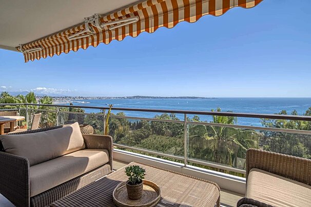 A sunny balcony with a striped awning, wicker furniture, and breathtaking ocean views of a distant coastline and lush greenery.