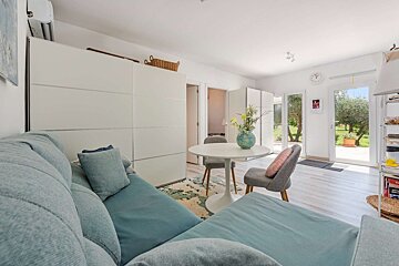 Bright living space featuring a blue sofa, white round dining table with flowers, and glass doors offering a view of a sunny green garden.