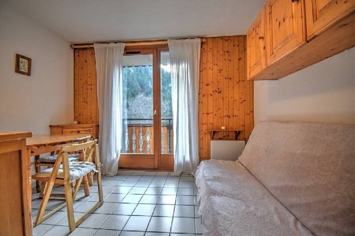 A cozy room features wood-paneled walls, a dining table, a sofa bed, tiled floor, and a balcony with a view of trees.