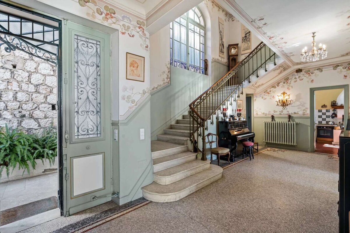 Elegant foyer with an elaborate staircase, floral murals, a piano, and an open ornate door leading to a stone courtyard.