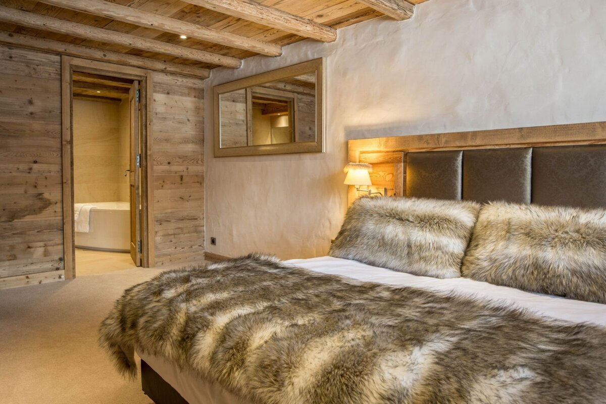 A cozy, rustic chalet bedroom features wood-paneled walls/ceiling, a bed with faux fur, and an open doorway to a bathroom.