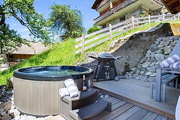An outdoor hot tub on a wooden deck with towels, wine, and a BBQ grill next to an alpine chalet.