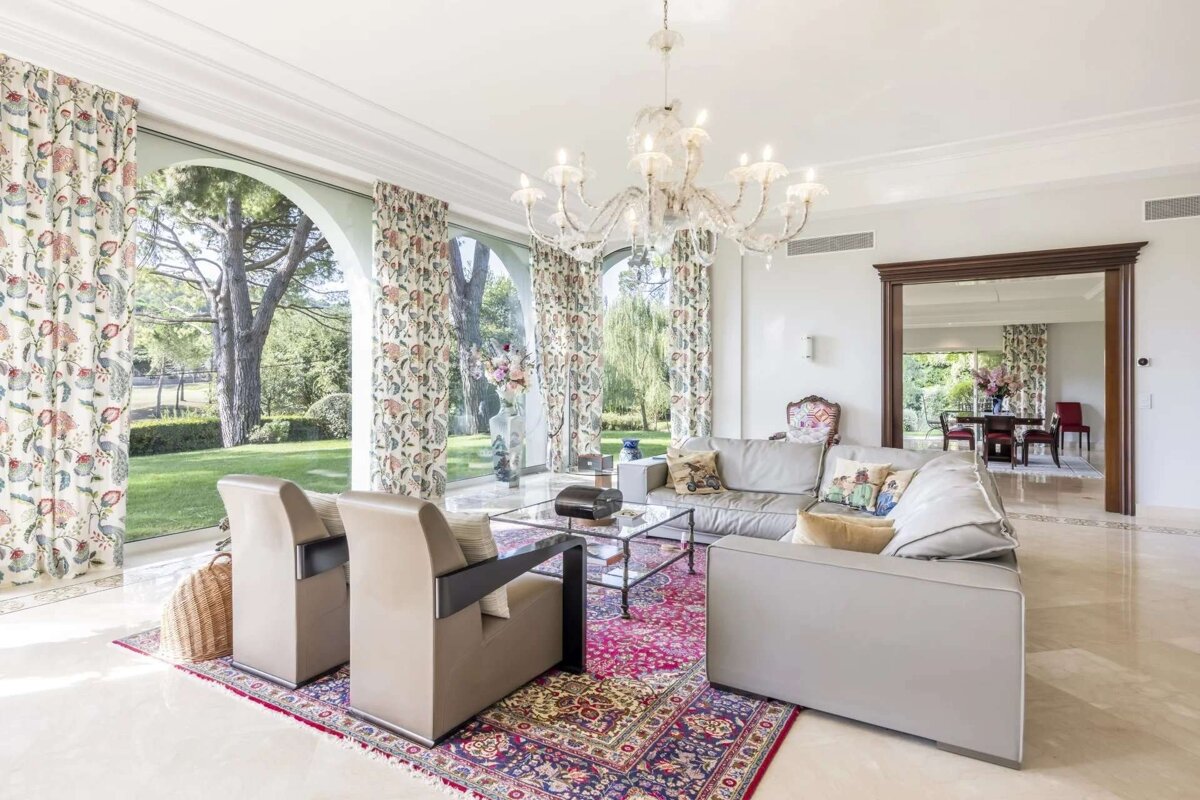 A bright, luxurious living room with large arched windows overlooking a lush garden. It features a grey sectional, elegant chandelier, and vibrant patterned rug.