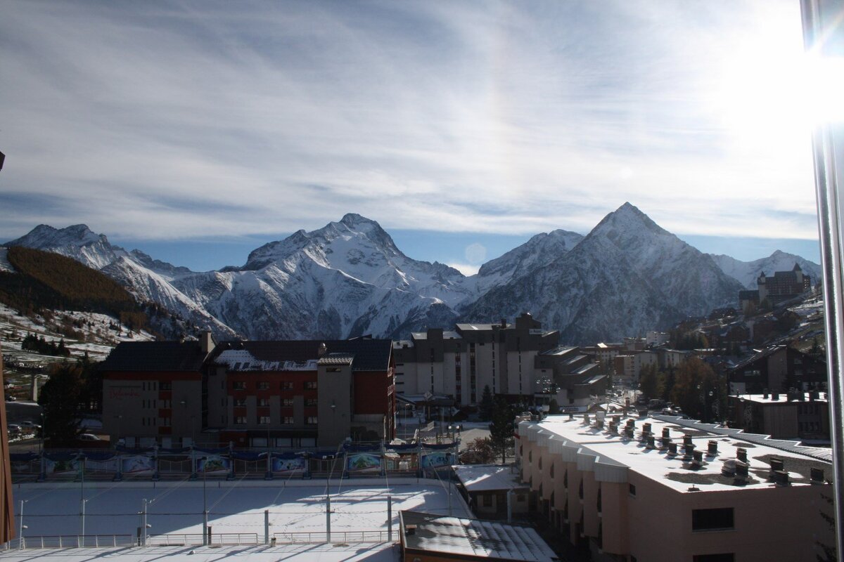 A snowy alpine town with an outdoor ice rink and buildings, set against majestic snow-capped mountains, under a bright sun.