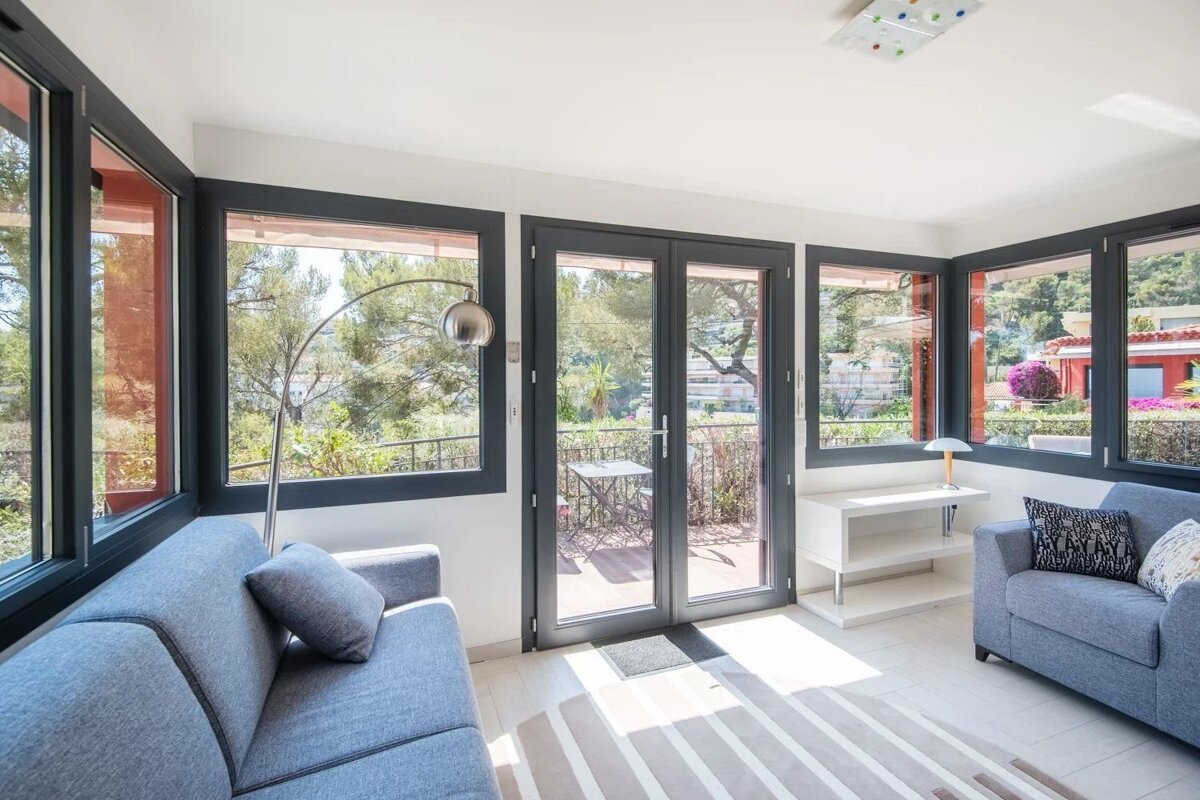 Bright modern living room featuring two blue sofas, large dark-framed windows, glass double doors to a balcony, and a striped rug.