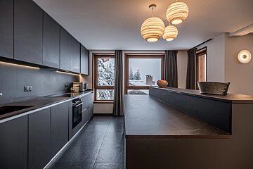 Modern dark kitchen with an island, warm pendant lights, and a snowy winter mountain view from large windows.