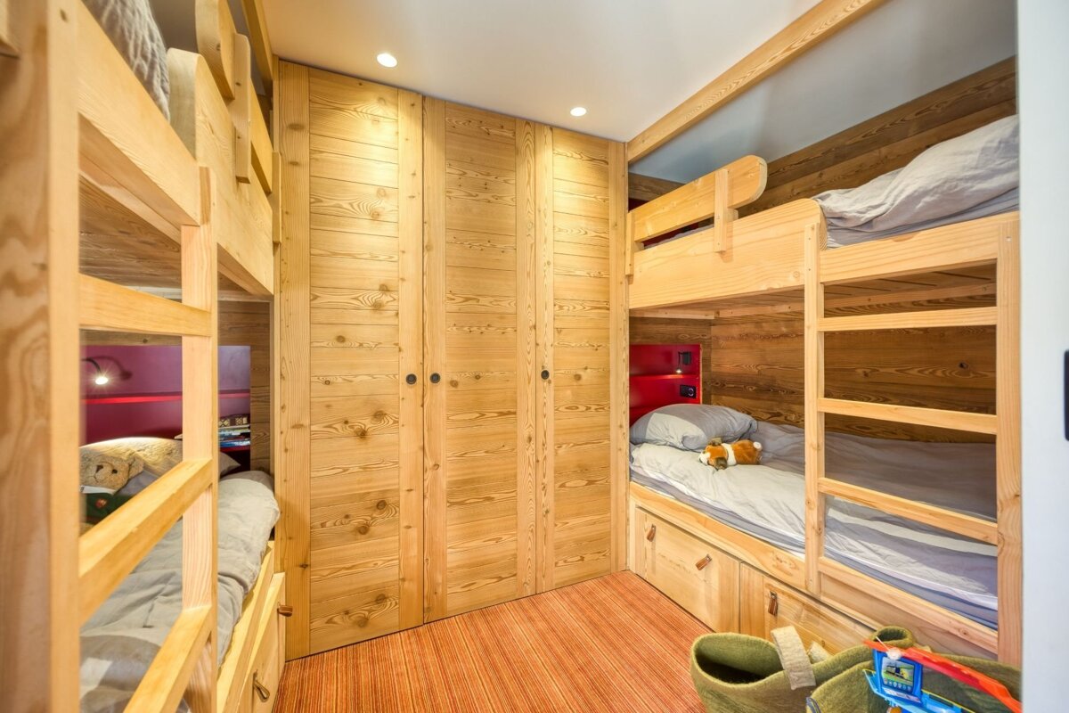A cozy, rustic bunk room features multiple light wood bunk beds, built-in wardrobes, and red accents, with an orange striped floor.