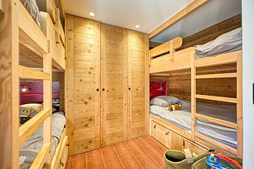 A cozy, rustic bunk room features multiple light wood bunk beds, built-in wardrobes, and red accents, with an orange striped floor.