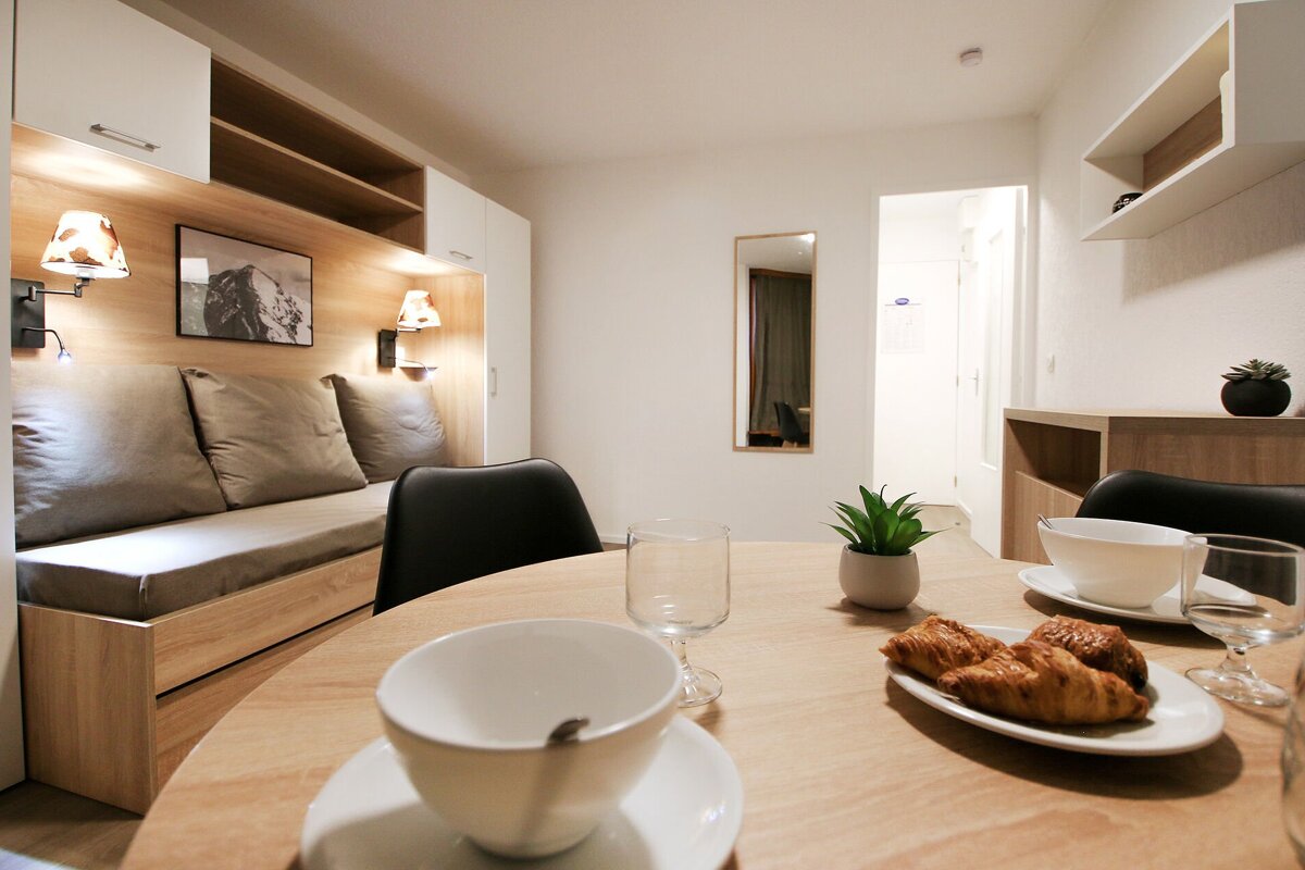 A bright, modern room features a wooden dining table with croissants, a built-in sofa bed, and light-toned, minimalist decor.