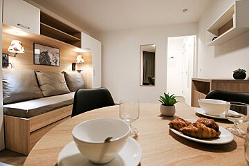 A bright, modern room features a wooden dining table with croissants, a built-in sofa bed, and light-toned, minimalist decor.