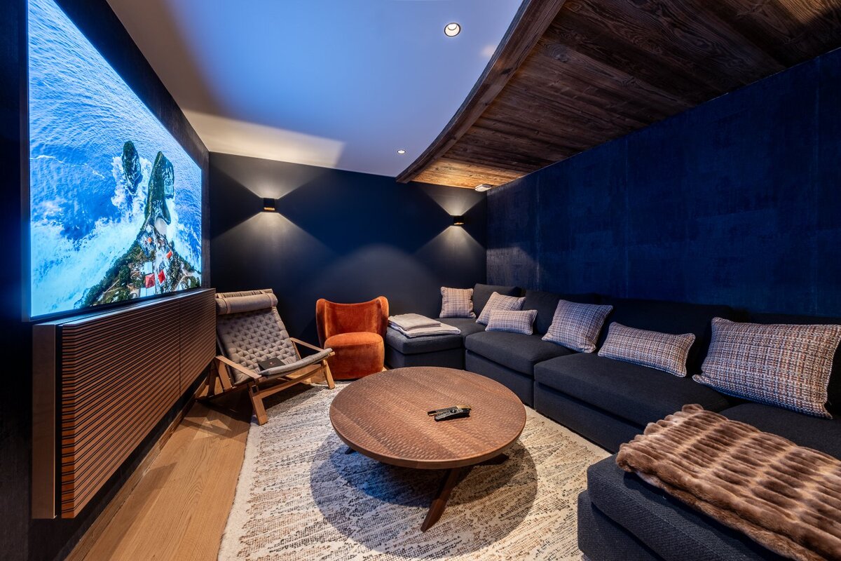 A modern, dark blue media room featuring a large TV, comfortable sectional sofa, and accent chairs, centered around a round coffee table.