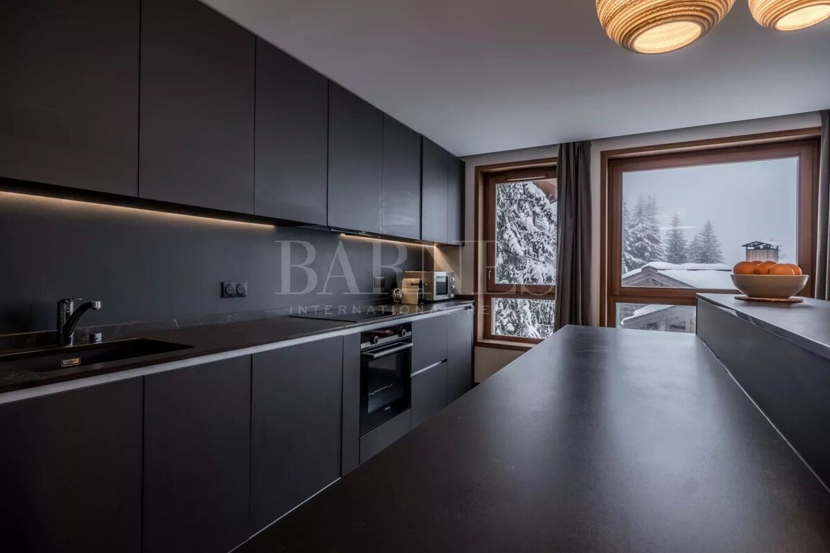 A modern, dark-themed kitchen with under-cabinet lighting. Large wooden windows reveal a serene snowy mountain landscape outside.