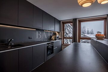 A modern dark kitchen with under-cabinet lighting, sleek black cabinets, and a snowy winter landscape seen through large windows. Oranges on the counter.