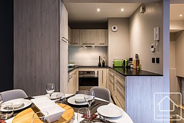 A modern, compact kitchen with light wood cabinets, black countertops, and a set dining table. Bright green appliances add contrast.