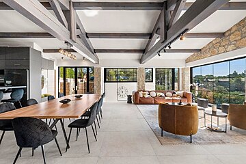Spacious open-plan living/dining room with rustic wood beams, stone accents, modern furniture, and large windows overlooking a green landscape.