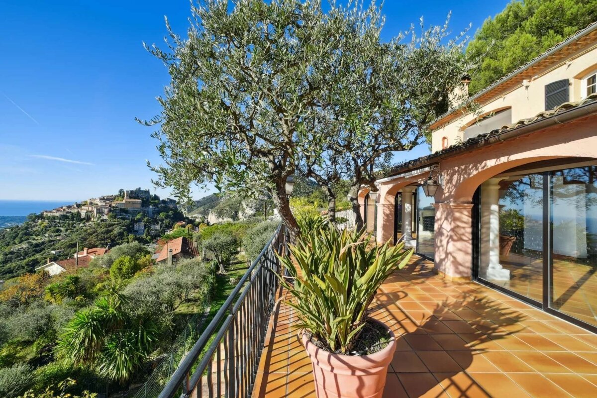 A sunny Mediterranean villa terrace with olive trees, offering a stunning panoramic view of a picturesque hillside village and the sparkling blue sea.