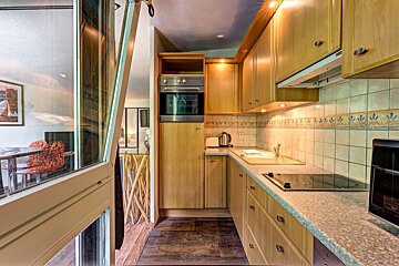 A narrow kitchen featuring light wood cabinets, tiled walls, built-in appliances, and a view into an adjacent room.