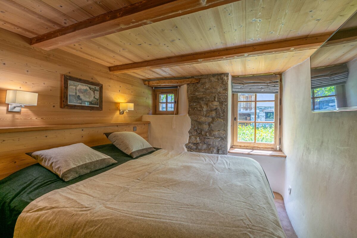 A warm, rustic bedroom with wood-paneled walls, exposed beams, and a cozy bed. A stone accent wall features windows letting in light.