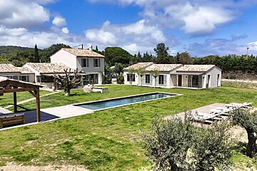 A sun-drenched property with traditional white houses, terracotta roofs, a long swimming pool, and sprawling green lawns under a blue sky.