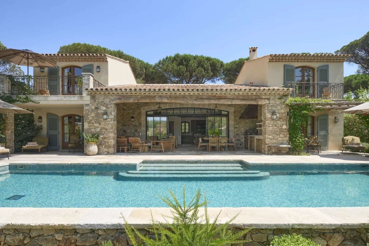 A luxurious stone villa with a sparkling swimming pool and expansive outdoor living spaces under a clear blue sky, evoking a Mediterranean feel.
