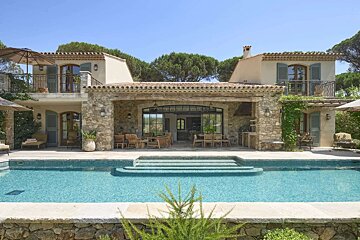 A luxurious stone villa with a sparkling swimming pool and expansive outdoor living spaces under a clear blue sky, evoking a Mediterranean feel.