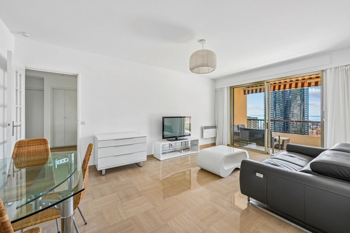 Bright, modern living room with a dark sofa, dining table, TV, and a balcony offering a city skyline and ocean view.