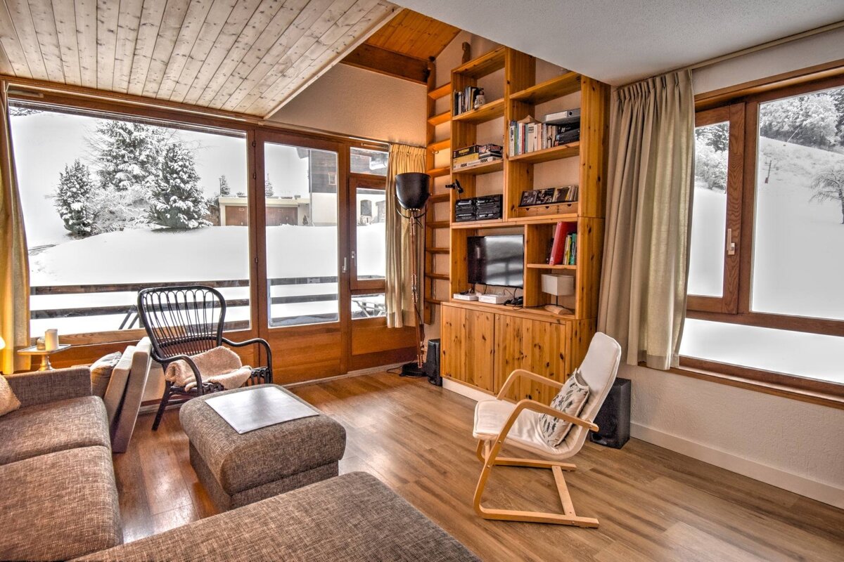 A cozy living room with wood accents, a sofa, bookshelves, and large windows overlooking a snow-covered winter wonderland.