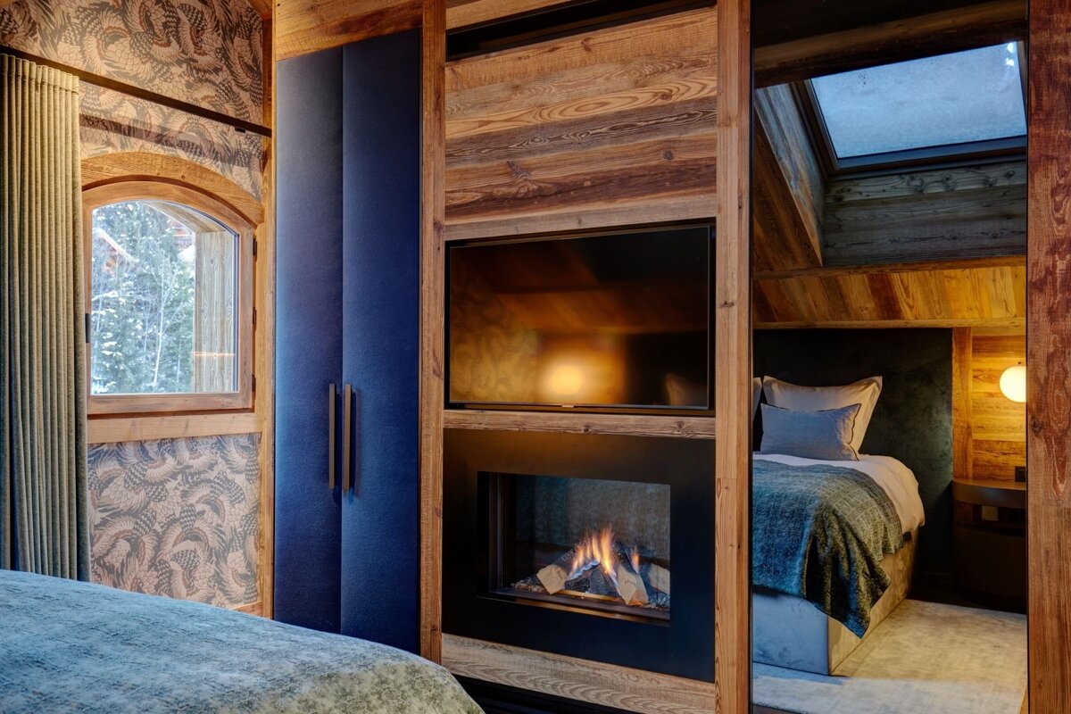 A warm, modern chalet bedroom featuring rustic wood, a cozy bed, fireplace, and TV. Blue accents complement the snowy window view.