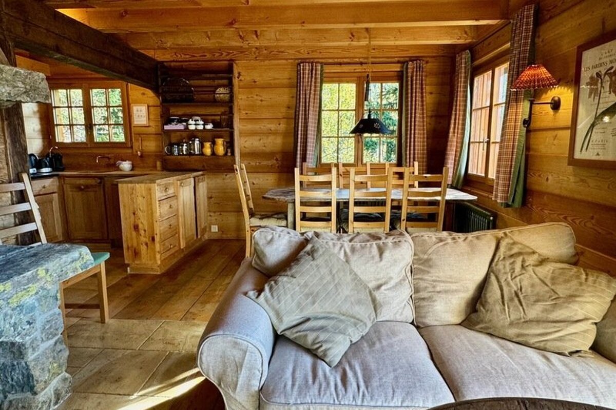 Cozy interior of a rustic wooden cabin featuring a comfy beige sofa, wood paneled walls, a dining set, and a kitchen corner.