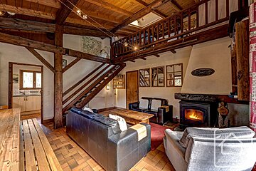 Cozy, rustic living room featuring exposed wooden beams, a warm fireplace, leather sofas, and a mezzanine. Terracotta floors complete the chalet feel.